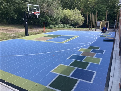 Outdoor residential sports court with a blue, green, and yellow surface, featuring a basketball hoop and hopscotch, surrounded by trees and greenery.