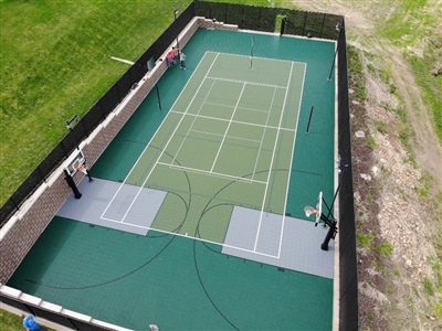 Outdoor sports court with a green and gray surface, featuring tennis nets and basketball hoops, surrounded by a black fence.