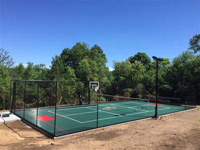 A green outdoor sport court with a basketball hoop and surrounded by fencing.