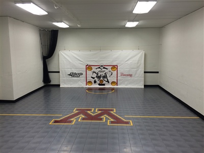 Indoor hockey practice area with grey flooring and a central U of M logo. 
