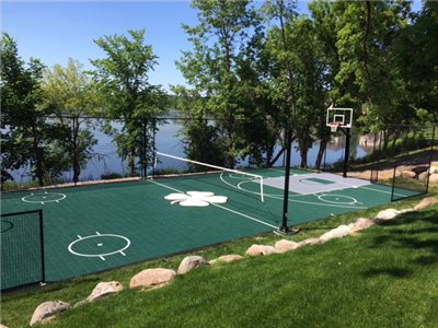 Outdoor multi-sport court with a green court and white central shamrock logo, with a volleyball net and a basketball hoop in Randolph.