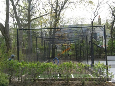 Multiple outdoor batting cages with black fencing next to a basketball court with grey flooring surrounded by greenery.