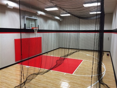 Indoor basketball court with wooden floor, red accent flooring, red safety padding below a basketball hoop and black netting in Afton.