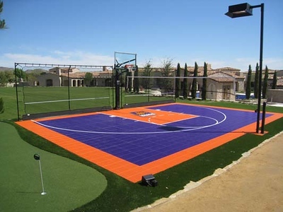 Outdoor residential basketball court with a purple and orange surface, featuring a basketball hoop and central logo, surrounded by a manicured lawn and a putting green.