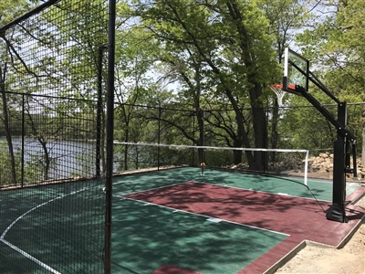 Outdoor court with a green and red surface, basketball hoop, and pickleball net, enclosed by fencing and surrounded by trees near a lake.