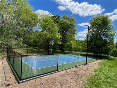 Outdoor fenced court with a blue surface and green border, featuring a basketball hoop and surrounded by trees and grass.