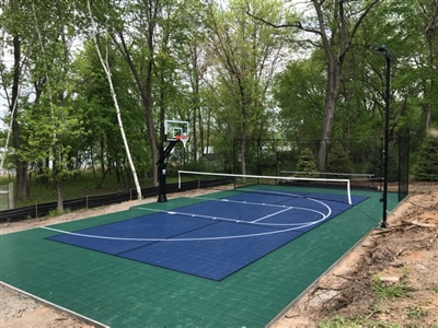 Outdoor court with a blue playing surface and green border, featuring a basketball hoop and pickleball net, surrounded by trees.
