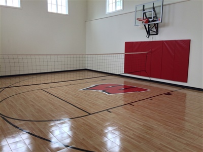 Indoor basketball and volleyball court with a polished wooden floor, featuring a red logo in the center, red padding on one wall, and a basketball hoop.