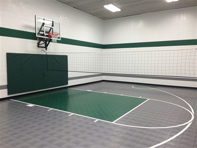 Indoor basketball court with a gray floor and green key area, featuring green wall padding and white walls with green accents, and a basketball hoop.