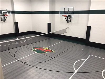 Indoor basketball court with a gray patterned floor, a green and red logo at center court, featuring two basketball hoops and a white volleyball net.