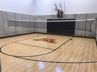 Indoor basketball court with a light wooden floor, featuring a red and yellow logo at center court, black wall padding, and featuring a basketball hoop and volleyball net.