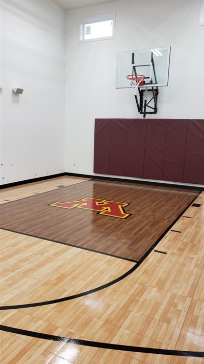 A small indoor basketball court with a polished wood surface, black lines, and white walls, featuring a U of M central emblem on a darker wood section, and a basketball hoop with maroon padding behind it in Minneapolis.