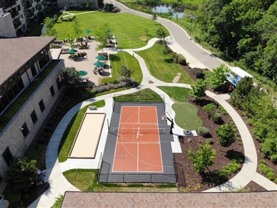 Outdoor residential sports area with an orange and black court, a sand volleyball court, and a putting green, surrounded by landscaped paths and greenery.