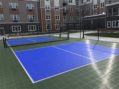 Outdoor sports court with a green surface and blue play areas, white lines, and tennis nets, surrounded by residential buildings in Blaine.