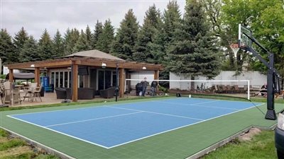 Outdoor residential sports court with a blue and green surface, a basketball hoop, and a nearby wooden pergola, surrounded by trees and greenery in Forest Lake. 