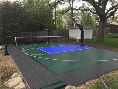 Outdoor residential sports court with a green and black surface, a blue painted key area, a basketball hoop, a volleyball net, and surround by a wooden fence. 