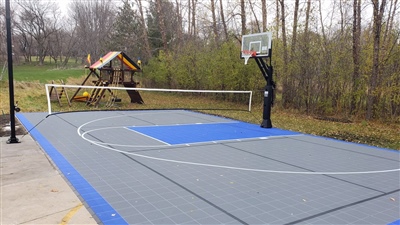 Outdoor residential sports court with a gray and blue surface, featuring a basketball hoop and a volleyball net.