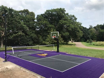  Outdoor residential sports court with a gray and purple surface, featuring a basketball hoop and a tennis net, surrounded by trees and greenery.